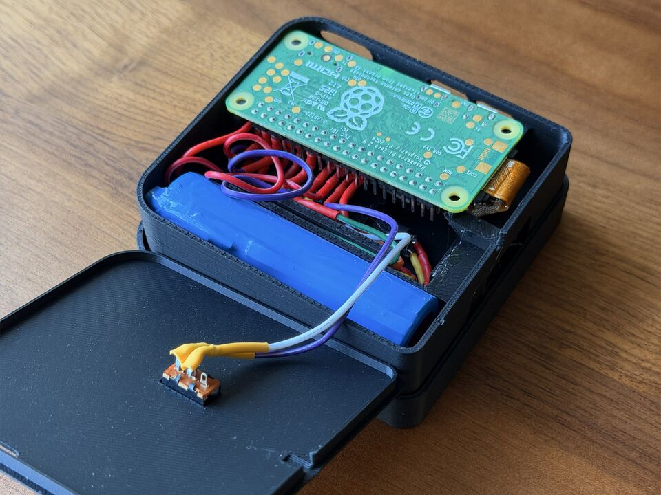 The cartridge with its lid lifted: the Raspberry Pi Zero 2 W on top, a blue 18650 cell beside it, red and purple wires to the header, and the slide switch soldered to the inside of the lid.